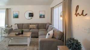 Un salon moderne avec un canapé d’angle, une table basse et une petite table d’appoint. Deux tableaux encadrés ornent le mur, ainsi qu’un miroir rond et un panneau mural sur lequel est écrit le mot « Salut ».