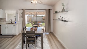 Une cuisine avec des meubles blancs, une table à manger avec des chaises, un ventilateur de plafond et une vue sur l’extérieur.