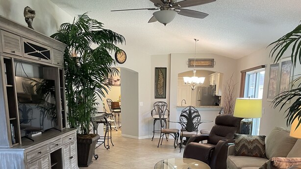 A living room with a sofa, armchairs, a coffee table, and a television. There is a ceiling fan, a potted plant, and a rug with a leaf pattern.