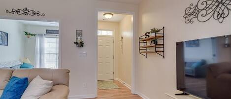 A living room with a beige sofa, a flat-screen TV on a stand, and a hallway leading to a door.