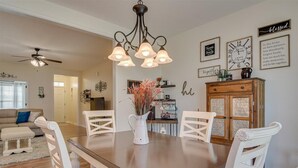 A dining area with a wooden table, white chairs, a chandelier, and a vase with dried flowers.