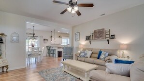 A spacious living room with a beige sofa, a matching bench, and a patterned rug. A ceiling fan and pendant lights are visible.