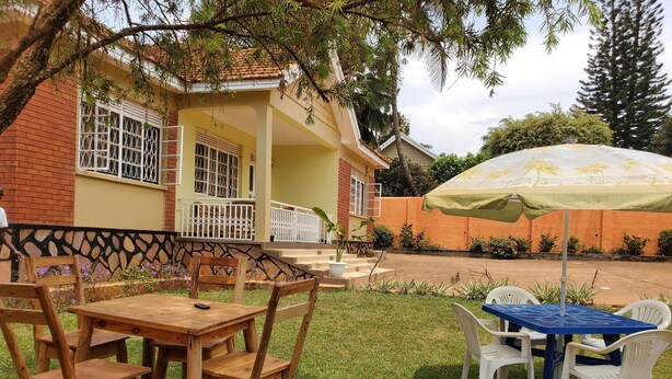 Une terrasse avec des tables et des chaises en bois et en plastique, un parasol vert et une maison en briques dotée d’un balcon.