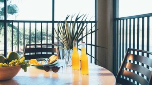 A table with a bowl of fruit, a glass vase with a plant, and two bottles of orange juice on a balcony with a view of greenery.