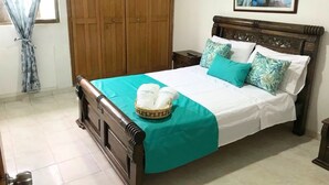 Een keurig opgemaakt bed met een blauwgroene sprei en kussens, een houten hoofdbord, en een mand met witte handdoeken aan het voeteneind.