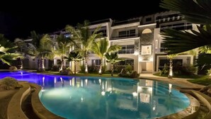 Vista nocturna de un complejo turístico con piscina, palmeras y un edificio moderno con varios balcones.