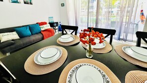 Un área de comedor con una mesa preparada para cuatro, un sofá con cojines y vista a una piscina por la ventana.