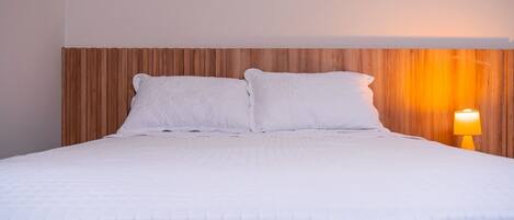 Una cama bien hecha con ropa blanca y almohadas, un cabecero de madera y una lámpara de noche.