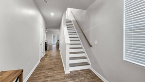 Un pasillo con suelo de madera, paredes blancas y una escalera con barandilla de madera.