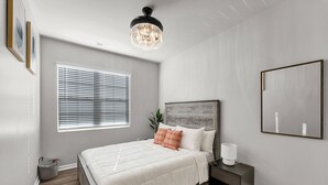Un dormitorio con una cama grande, una mesita de noche, una lámpara, una araña y una ventana con persianas, además de un cuadro en la pared.