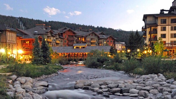 Ein Blick auf eine Lodge am Flussufer, mit mehreren Gebäuden, einer Steinbrücke und einem bewaldeten Hintergrund.