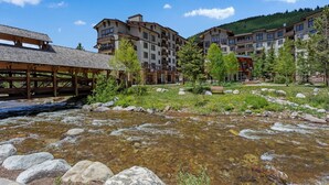 Aussicht auf einen Fluss mit einem Resort, mehreren mehrstöckigen Gebäuden, einer Holzbrücke und einem klaren Himmel.
