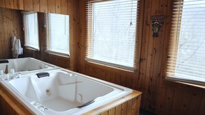 A wooden cabin bathroom with a large bathtub, wooden walls, and windows with blinds.