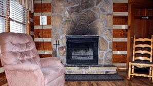 A stone fireplace with a log fire, a plush pink armchair, and a wooden chair in a room with wooden walls and flooring.