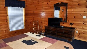 Ein Schlafzimmer einer Holzhütte mit Bett, Kommode, Fernseher und einem Fenster mit Jalousien.