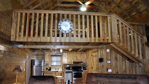 Interior rústico de una cabaña con techo de madera, un reloj central, zona de cocina y comedor.