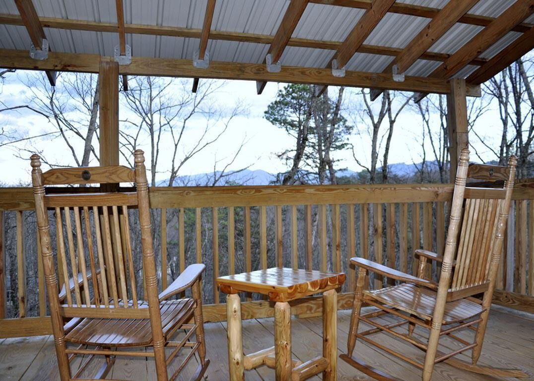 Terraza de madera con dos mecedoras y una pequeña mesa, con vistas a un paisaje montañoso.