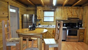 Interior de una cabaña de madera con una mesa redonda, sillas, un refrigerador, una estufa y armarios de madera.