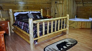 Una habitación de cabaña de madera con una cama, una mesita de noche, una lámpara, un banco de madera, una puerta de madera y un baño con lavamanos y bañera.