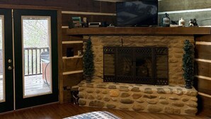 Ein gemütliches Zimmer mit Kamin aus Stein, einem Fernseher auf einem Holzboden und Blick durch eine Glastür.