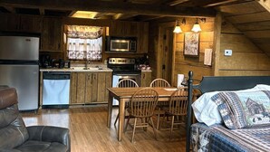 Ein gemütlicher Raum im Chalet-Stil mit Holzwänden, einem Esstisch mit Stühlen, einem Bett mit gemustertem Überwurf und einer Küchenzeile mit Kühlschrank, Mikrowelle und Herd.