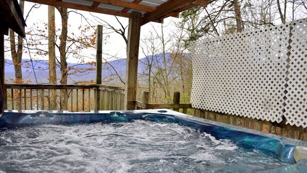 Ein Whirlpool mit einer Holzterrasse und einem weißen Gitterzaun, umgeben von Bäumen und Bergen im Hintergrund.