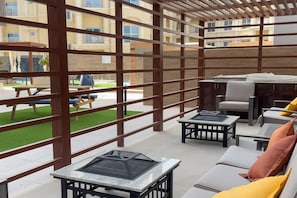 Ein überdachter Außenbereich mit Tischen und Lounge-Möbeln, einem Grill und Blick auf einen Innenhof mit Rasen und Gebäuden im Hintergrund.