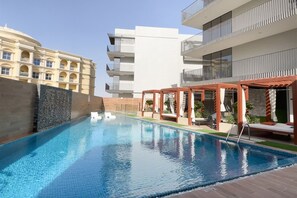 A swimming pool with a clear blue water surface, surrounded by a wooden deck and a stone wall. - Enchanting 1BDR Apt w/ Direct Pool & Gym Access (Arjan)