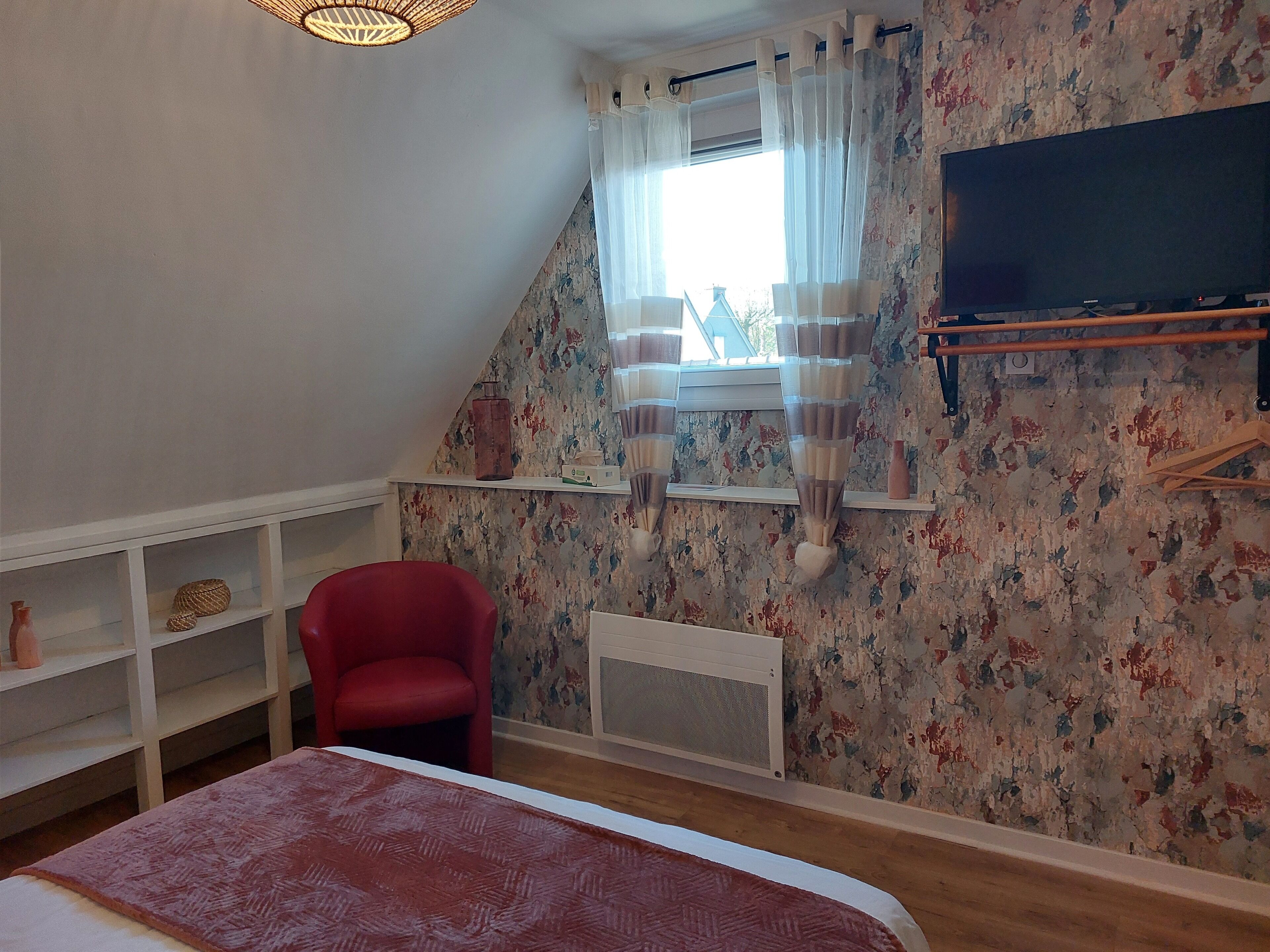 Een slaapkamer met een schuine balklaag, een rode stoel, een bed met een beddegoed met patroon, een radiator en een aan de muur bevestigde televisie.