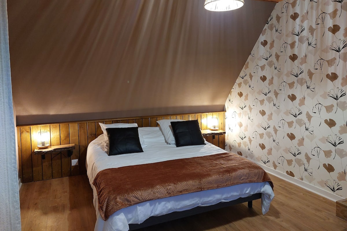 Ein Schlafzimmer mit schrägen Wänden, einem hölzernen Kopfteil, zwei Nachttischlampen, einem Bett mit weißer und brauner Bettwäsche und einem floralen Wandmuster.