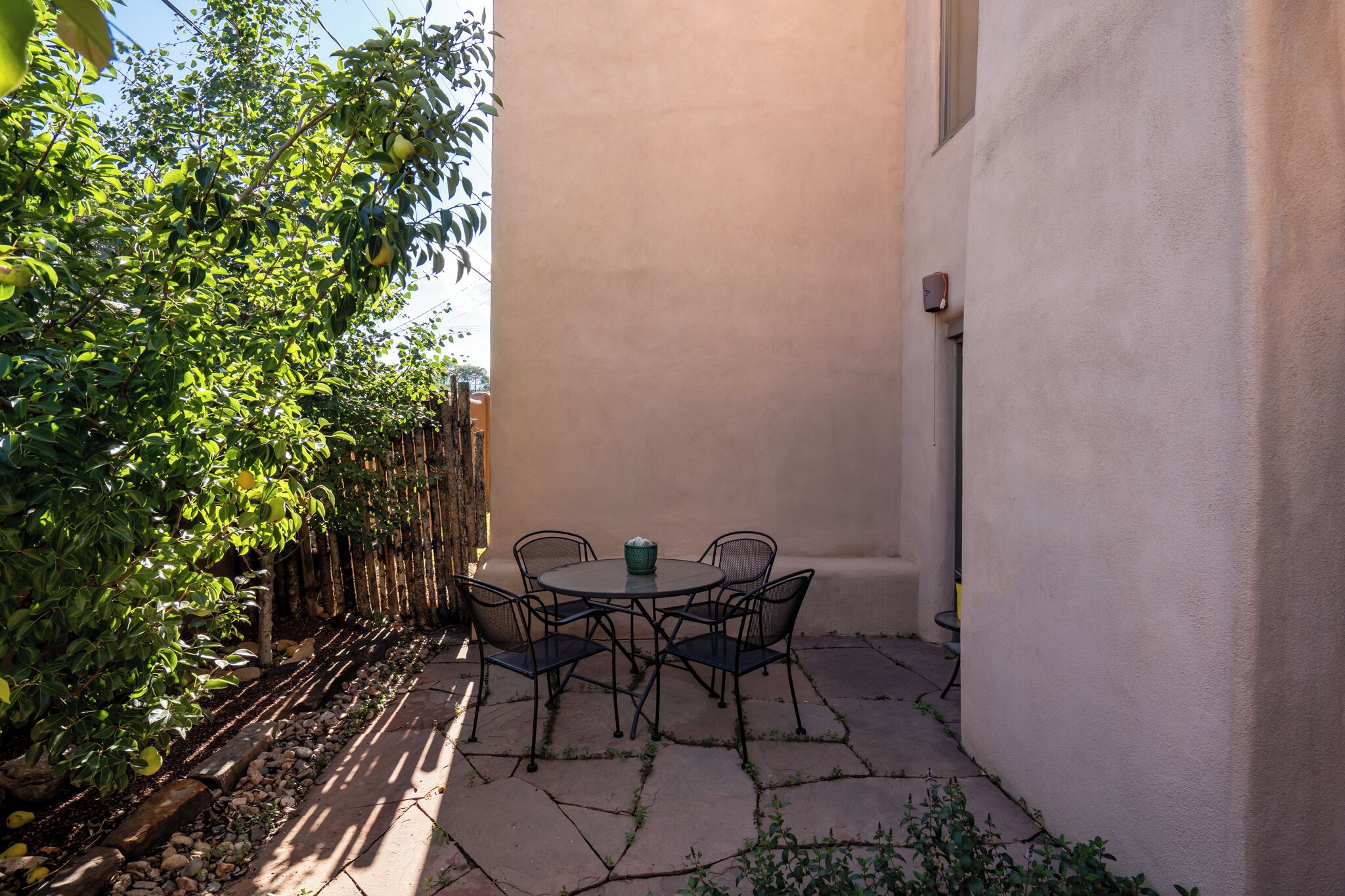 Un patio con una mesa redonda y sillas, rodeado por un bajo muro de piedra y abundante follaje.