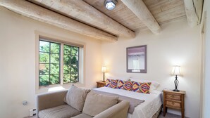 Un dormitorio con una cama grande, dos mesitas de noche con lámparas, una ventana con vistas a la vegetación, techo de madera y un sofá color beige.
