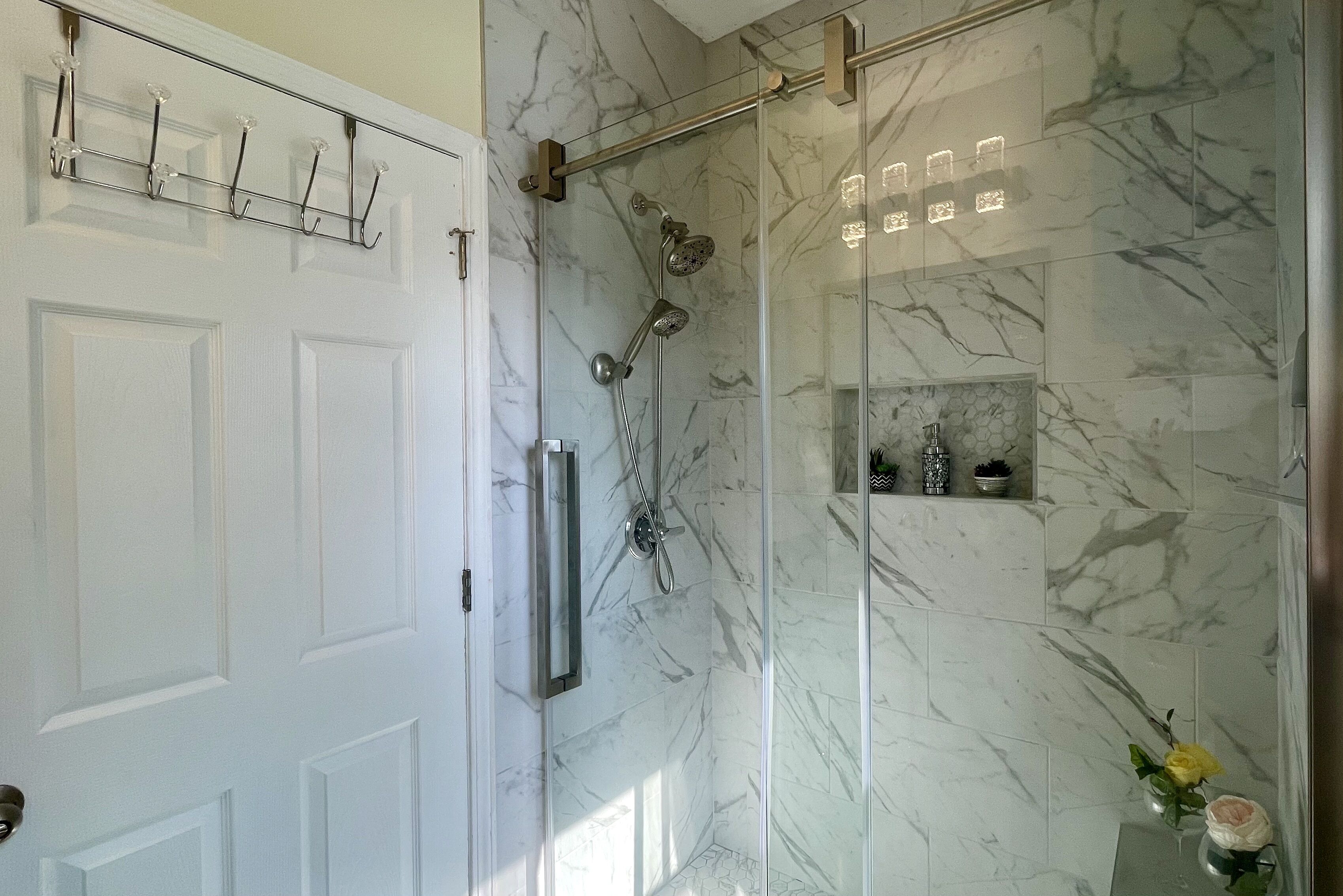 Baño con cabina de ducha de vidrio, baldosas de mármol, una puerta blanca y un pequeño estante con artículos de aseo.