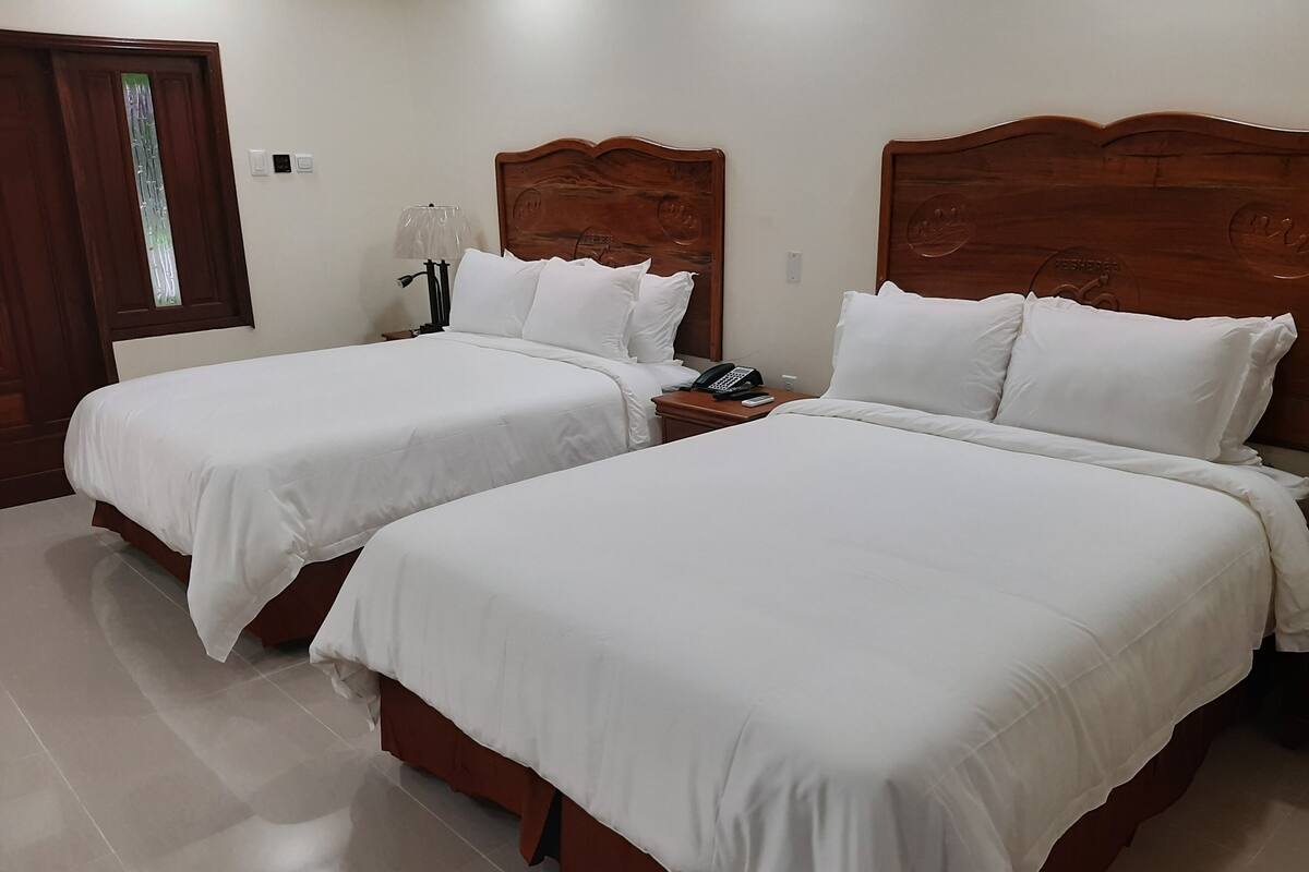 A hotel room with two beds, wooden headboards, white bedding, a bedside table with a lamp, and a phone.