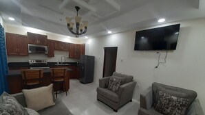 A modern living room with a grey sofa, a dining area with wooden chairs, a kitchen with wooden cabinets, a black refrigerator, and a flat-screen TV mounted on the wall.
