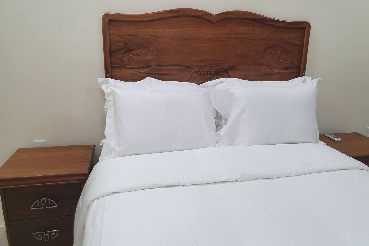 A neatly made bed with white bedding and pillows, a wooden headboard with carved details, and a wooden nightstand with drawers.