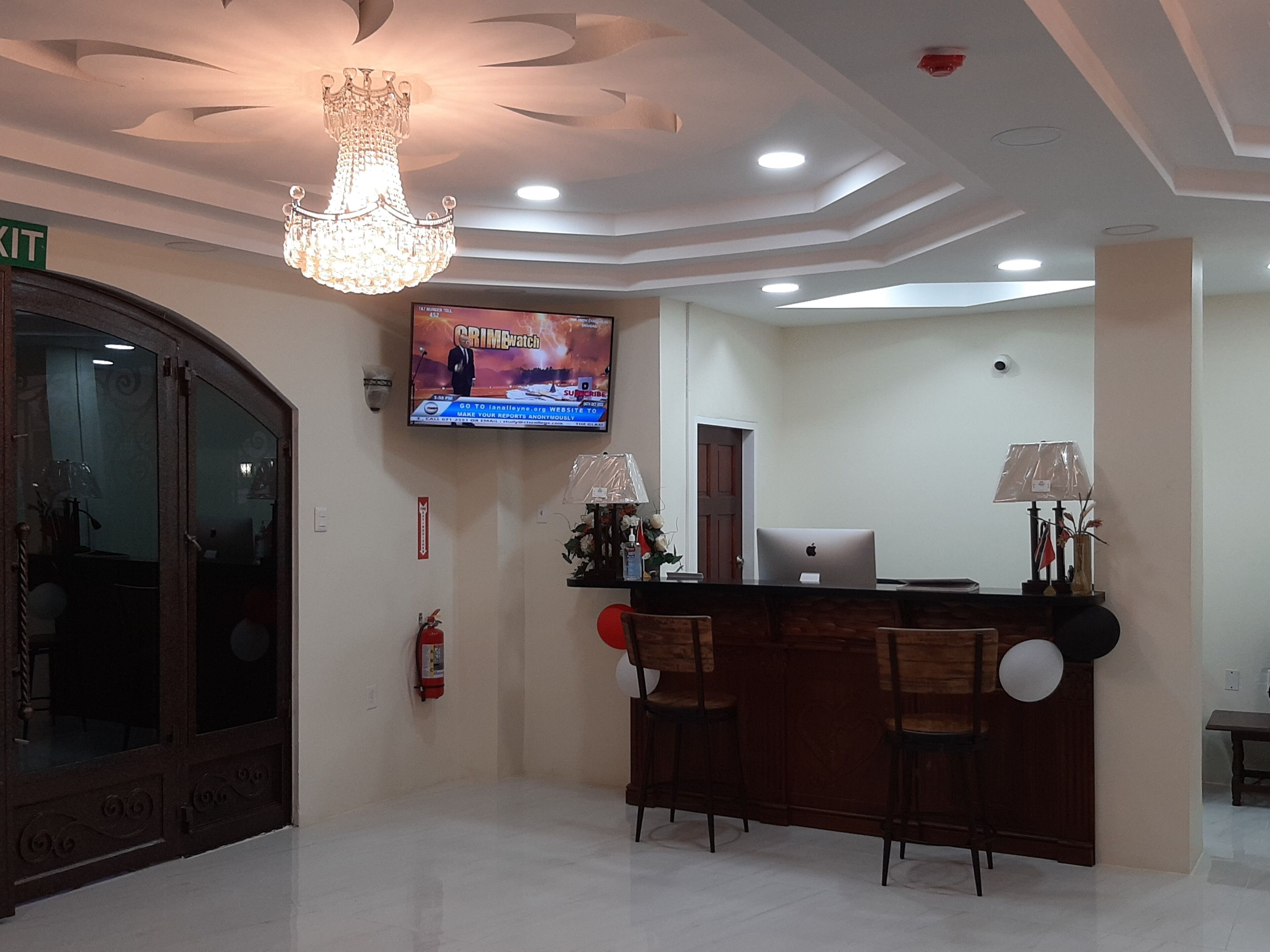 Un'area reception con un lampadario, una televisione che mostra 'Crime Watch' e una scrivania con sedie.