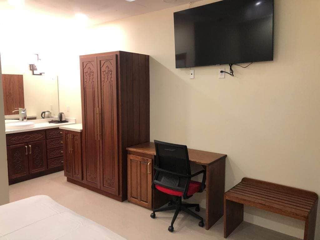 A bedroom with a bed, a desk with a chair, a TV mounted on the wall, and wooden cabinets.
