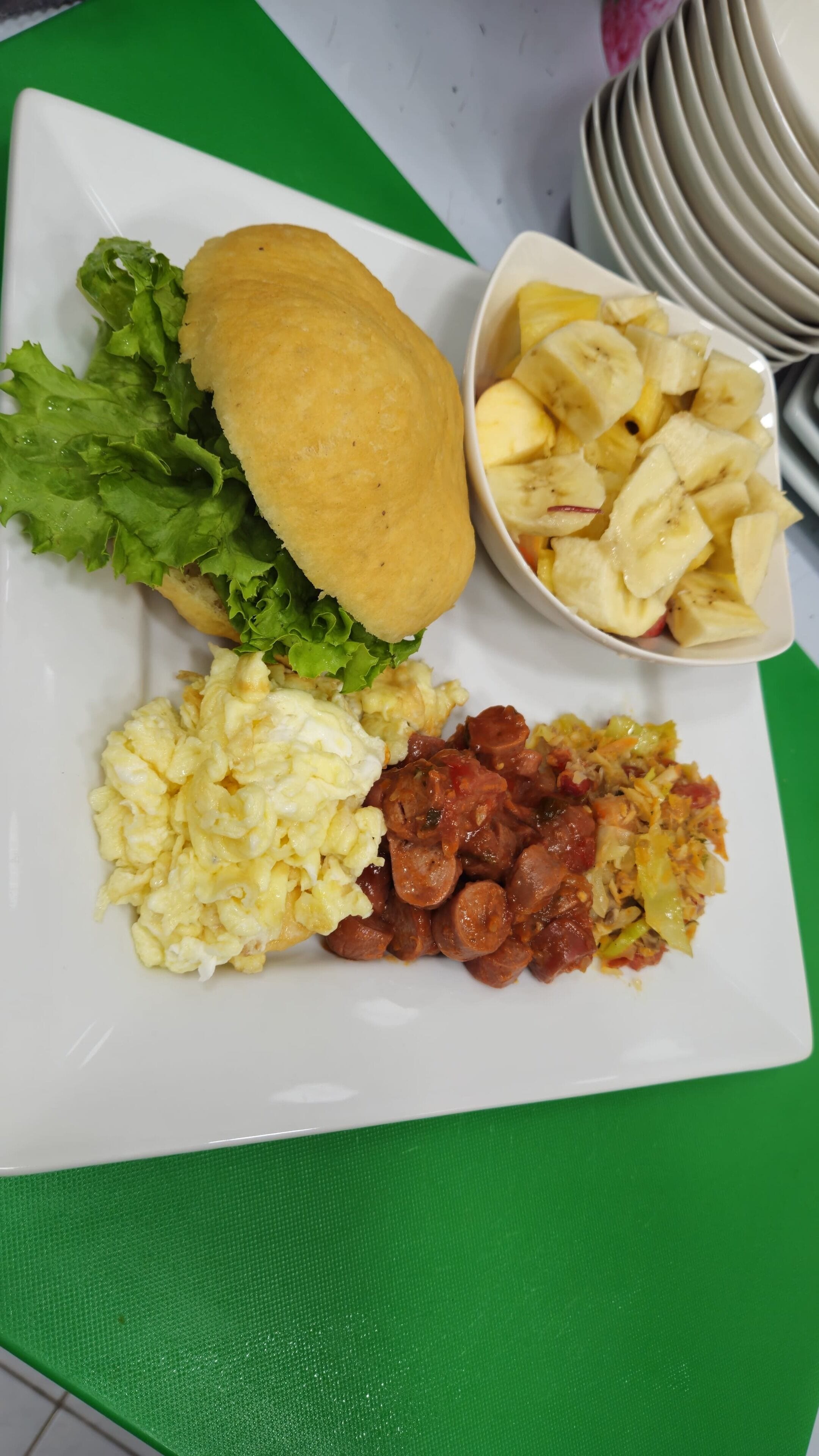 A plate with scrambled eggs, sausage, a sandwich, and a bowl of banana chips.