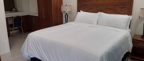 Una cama bien hecha con ropa de cama blanca, cabecera de madera y mesita de noche con lámpara.
