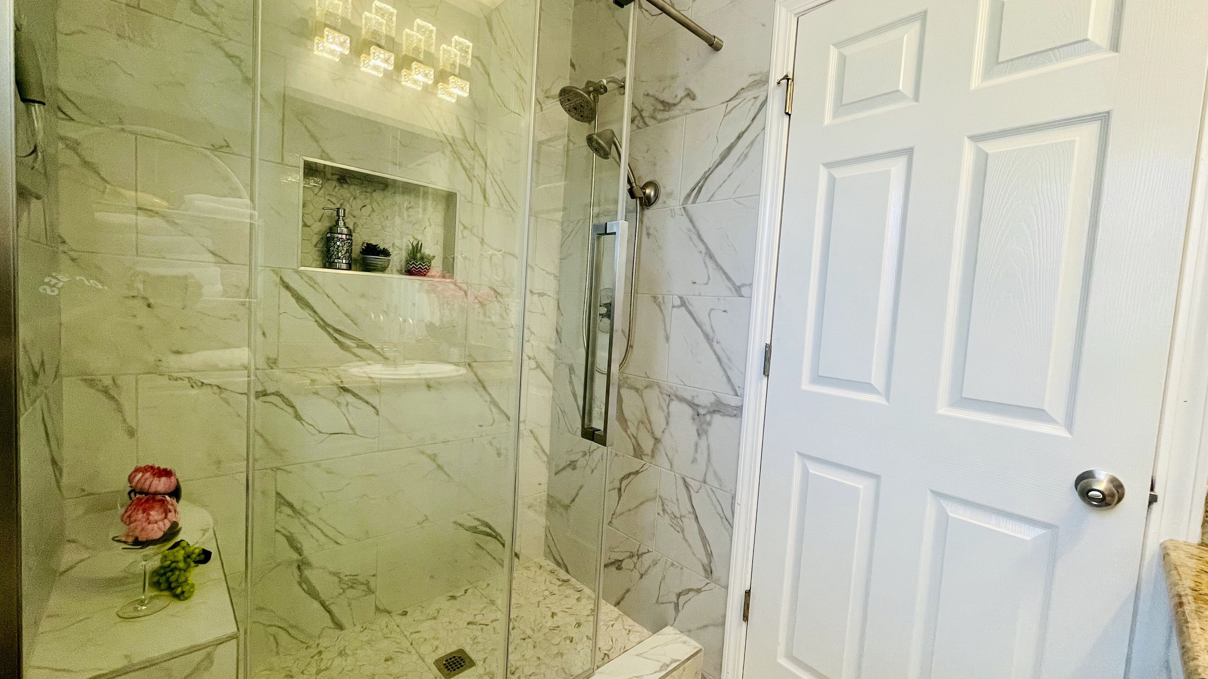 Une salle de bain avec une douche en marbre, un meuble-lavabo entouré de parois en verre et une porte blanche.