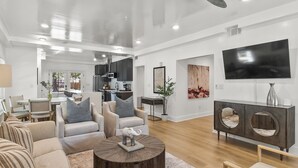 A spacious living room with a modern ceiling fan, a large flat-screen TV, and a dining area in the background.