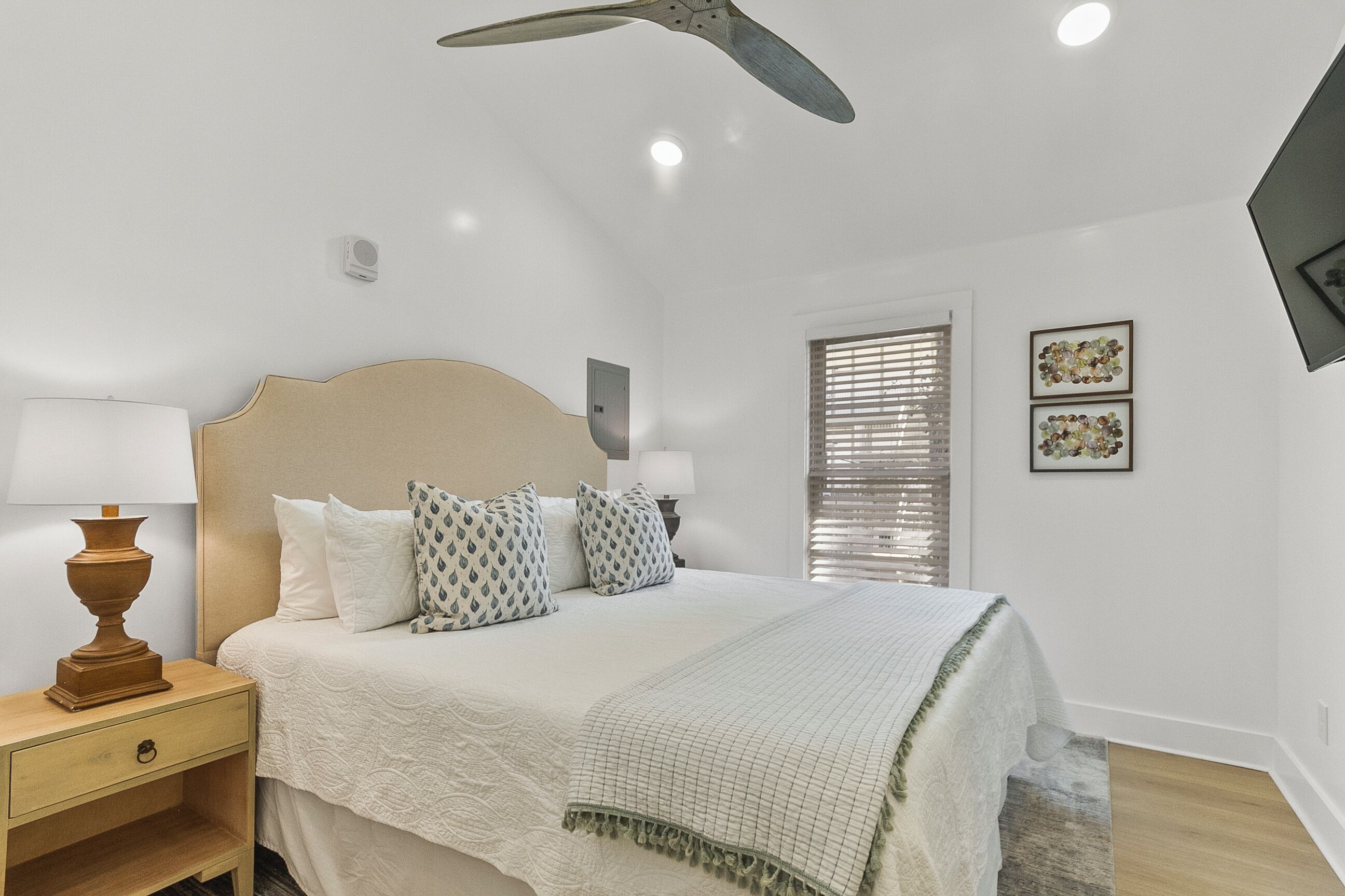 A bedroom with a bed, bedside tables, a ceiling fan, a window with blinds, and wall-mounted artwork.