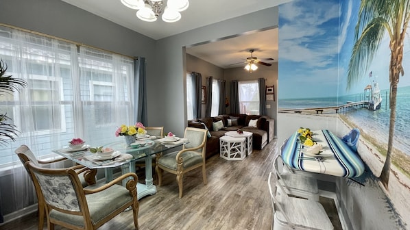 A dining area with a glass table, chairs, and a view of a beach mural.