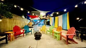 Un área de estar al aire libre con sillas Adirondack de colores dispuestas alrededor de un hogar para fuego central.