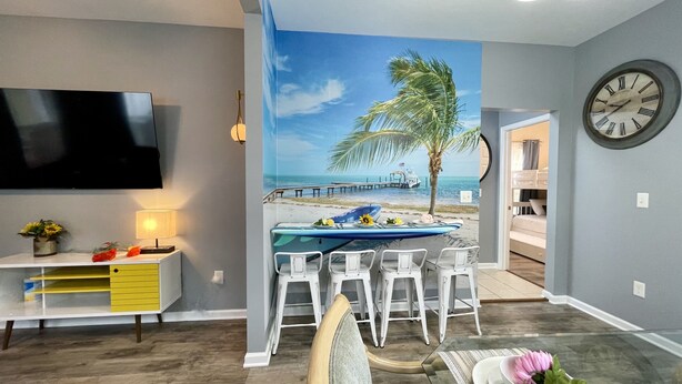 Una habitación con un mural de playa, un comedor con sillas blancas, un televisor montado en la pared y un reloj en la pared.