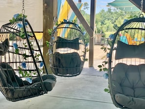 Two hanging chairs with cushions and blue flowers, under a shaded area with a view of greenery.