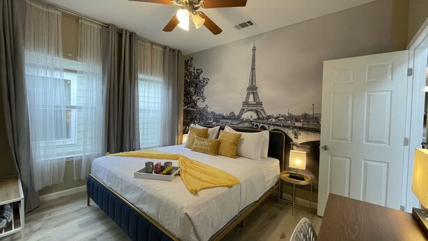 Ein Hotelzimmer mit Bett, Nachttisch, Lampe, einem Fenster mit Vorhängen und Wandbild von Eiffelturm.