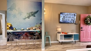 Una stanza con un affresco raffigurante una scena subacquea, una TV a schermo piatto, una porta rosa e un piccolo tavolo con una lampada e fiori.