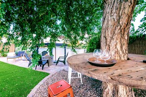 A wooden table with a glass and a tray, surrounded by chairs, under a tree.